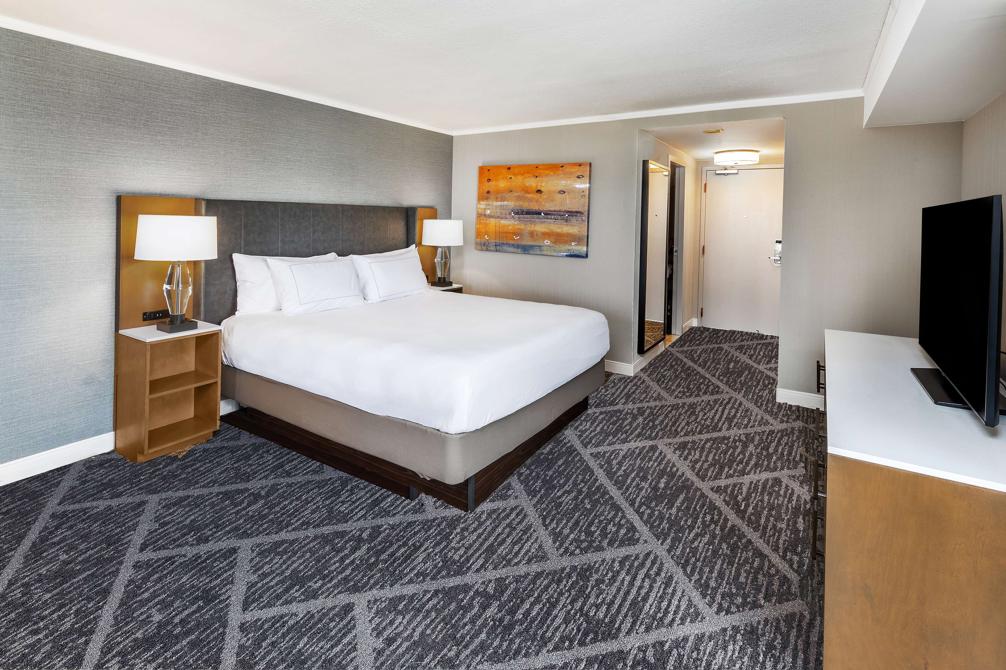 A hotel room with a large bed, a nightstand with a lamp, a television, and a carpeted floor.