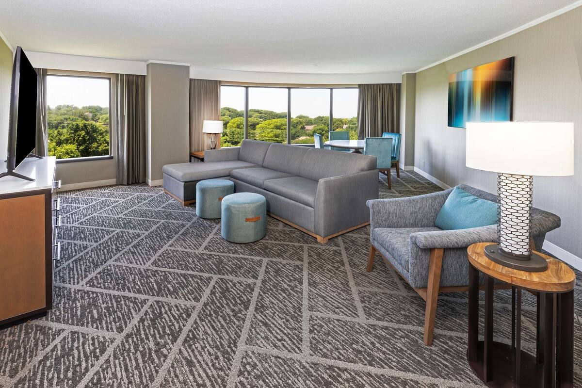 A hotel room with a large gray sectional sofa, a smaller gray armchair, a round wooden side table with a lamp, a flat-screen TV, and a dining table with chairs.