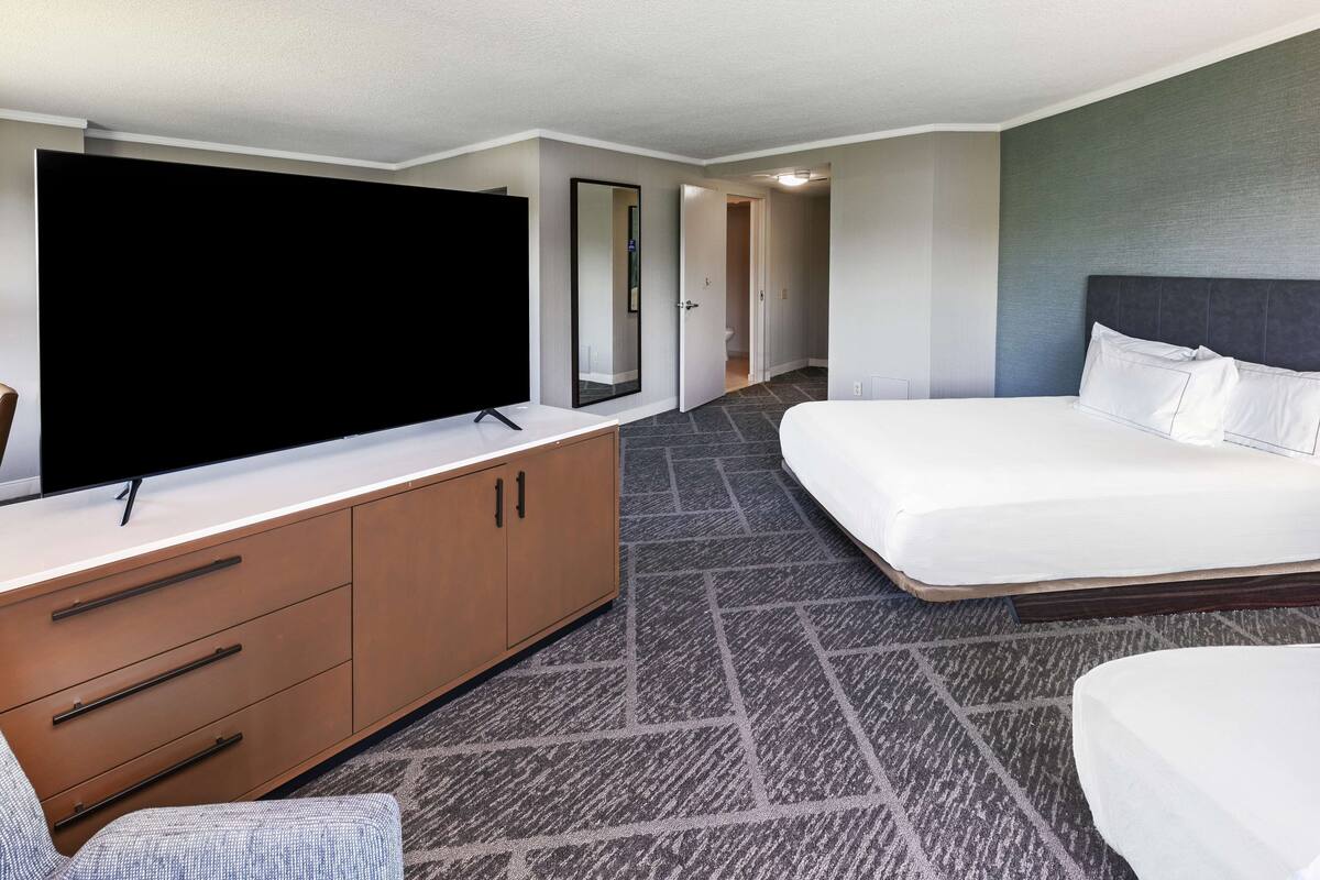 A hotel room with a large flat-screen TV, a bed with white linens, a wooden dresser with drawers, and a carpeted floor.