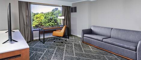 A hotel room with a gray sofa, a desk with a computer, a chair, and a large window offering a view of trees.