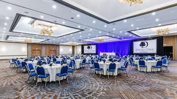 Una amplia sala de conferencias con mesas redondas y sillas azules, un escenario con cortina azul, y un cartel de 'DoubleTree by Hilton'.