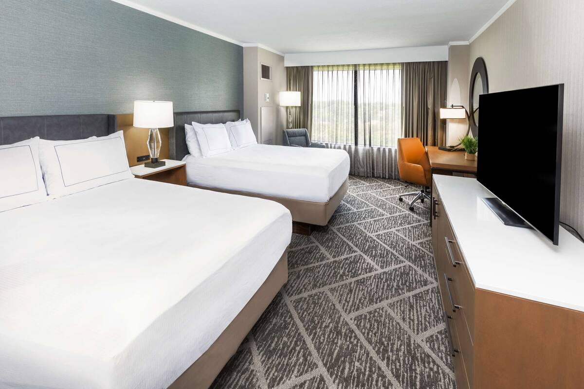 A hotel room with two beds, a desk with a TV, a chair, and a large window with curtains.