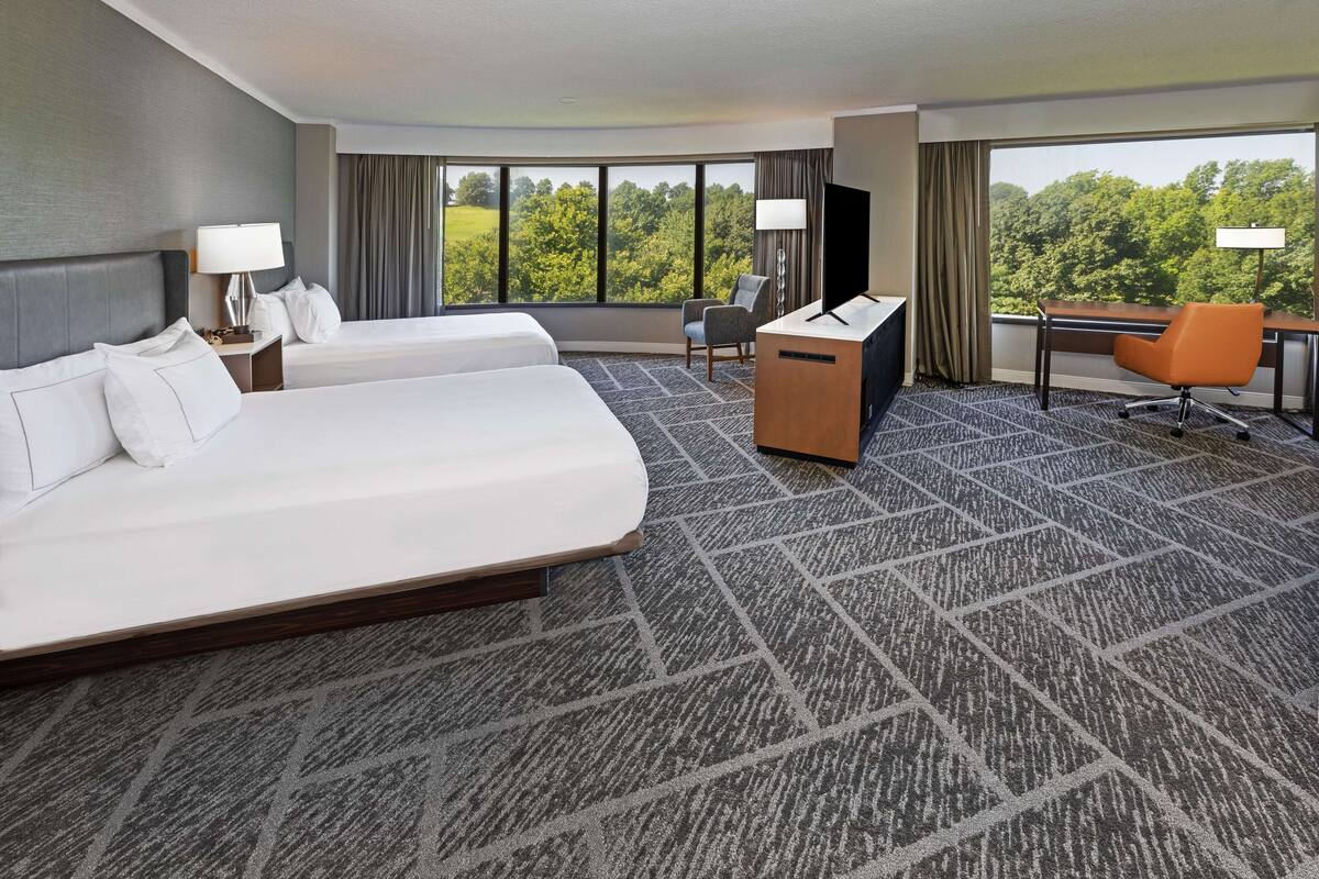 A hotel room with two beds, a desk, a chair, and a large window offering a view of trees.