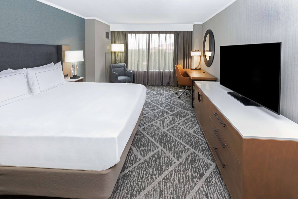 A hotel room with a large bed, a desk with a flat-screen TV, a chair, a desk lamp, and a window with curtains.