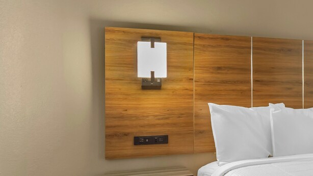 A hotel room with a wooden headboard, a bedside table, and white bedding. - Red Roof Inn Lumberton (Lumberton)