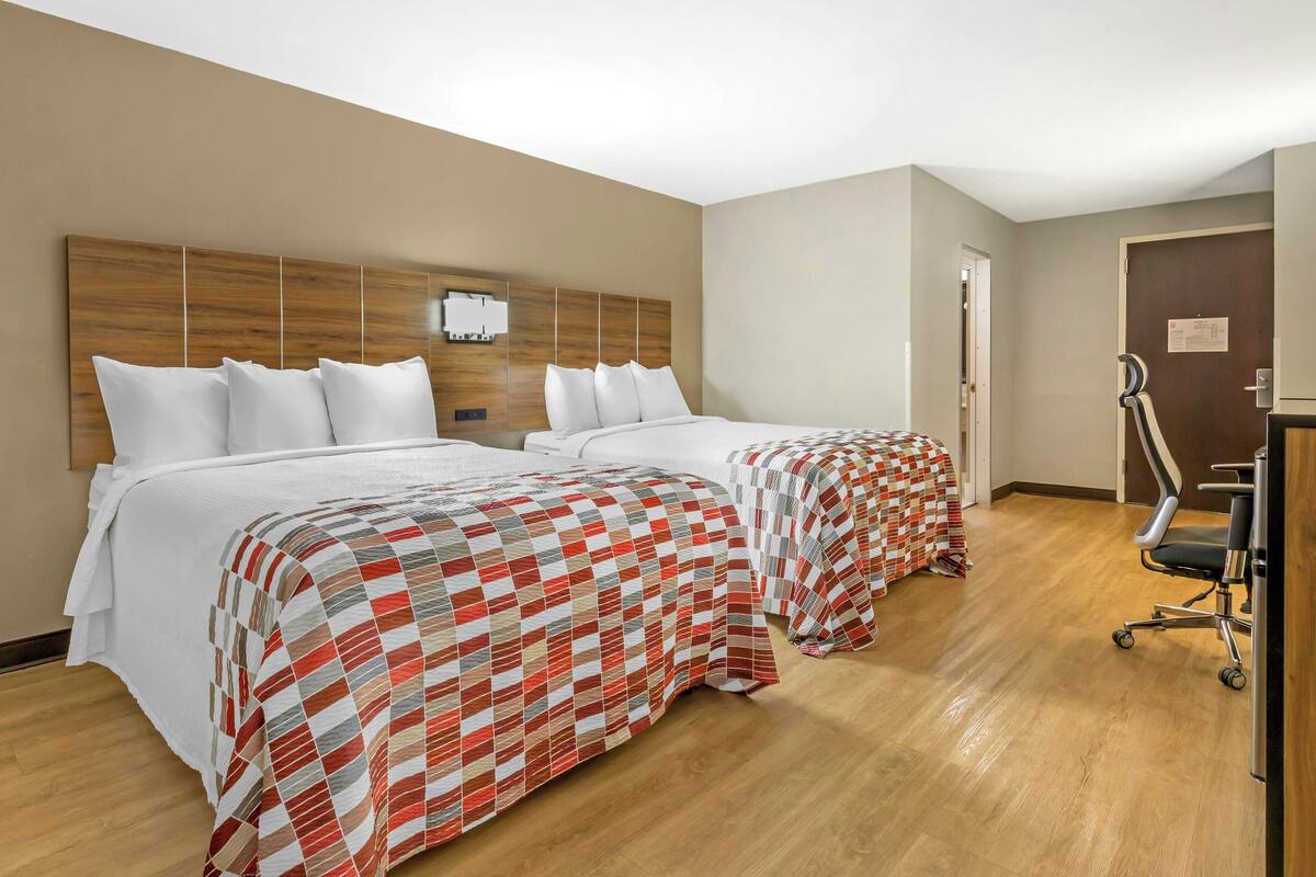 Une chambre d’hôtel avec deux lits, une tête de lit en bois, un bureau avec une chaise et une porte.