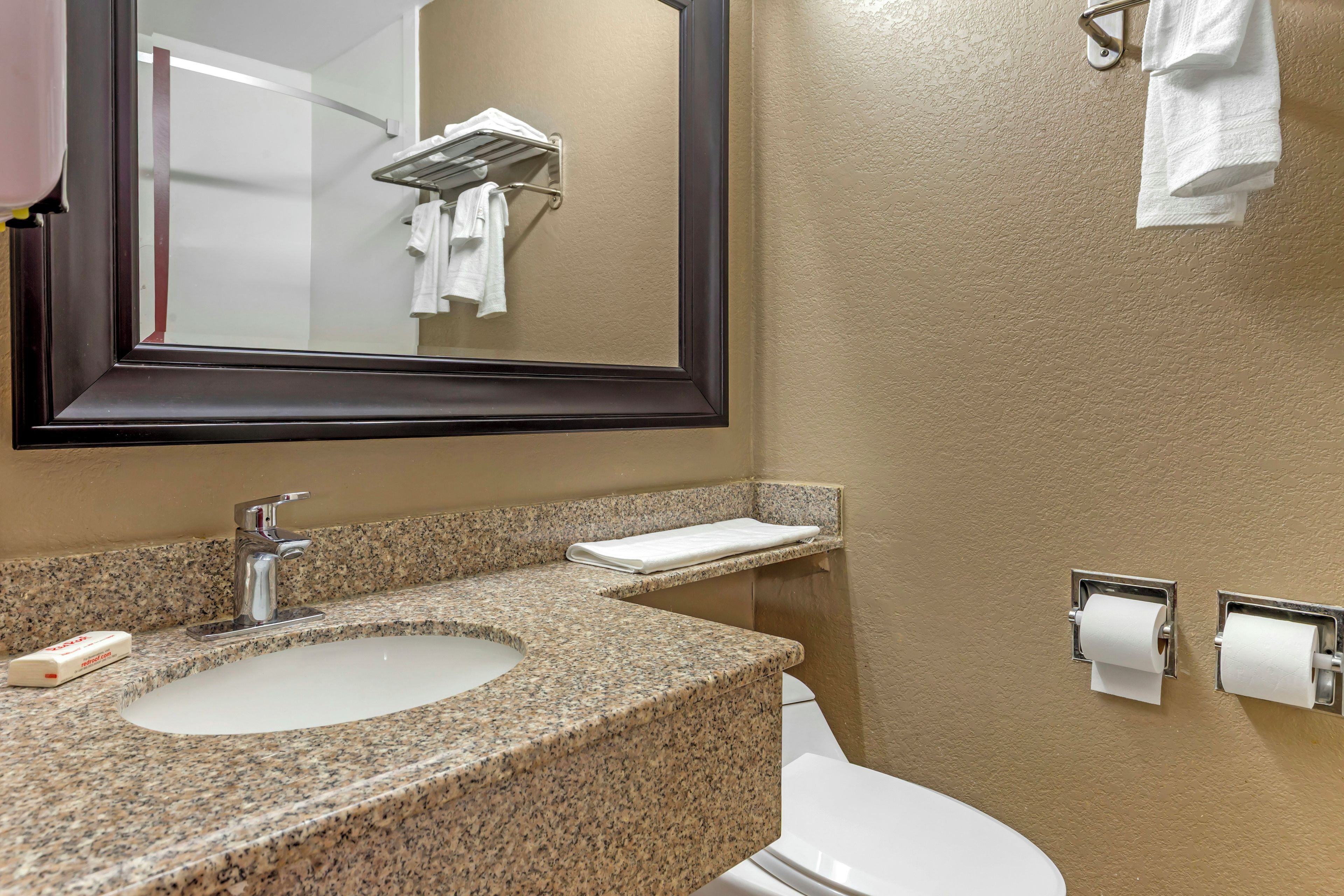 Une salle de bain avec un lavabo doté d’un plan de travail en granit, un miroir, un porte-serviettes et des toilettes avec des essuie-mains en papier.