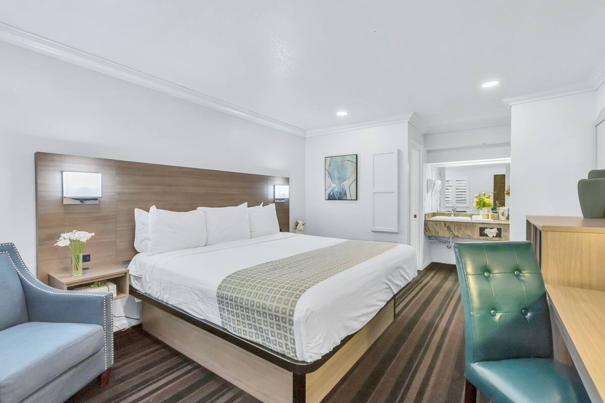 A hotel room with a large bed, a desk, a chair, a vanity with a mirror, and a small table with flowers.