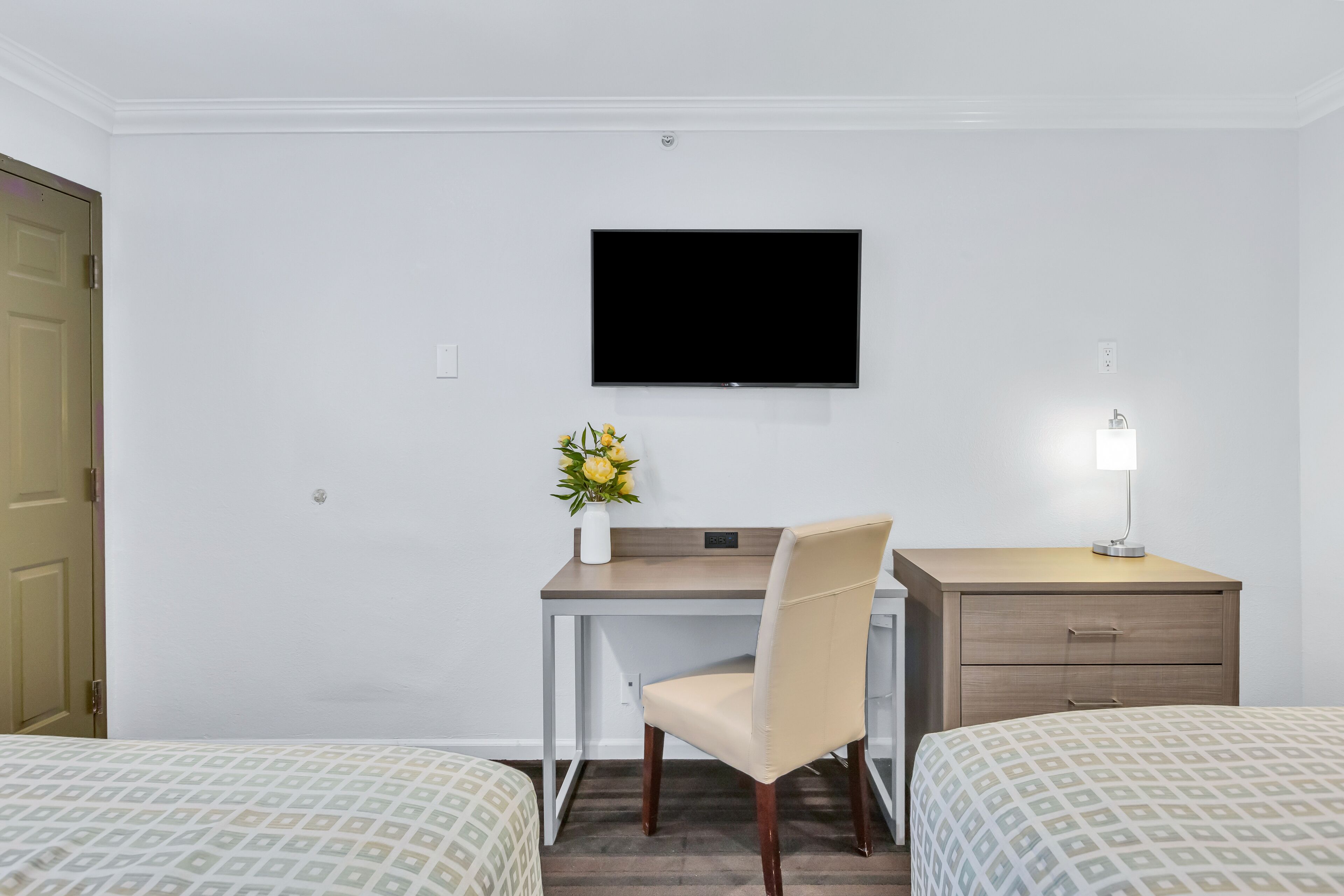 A hotel room with a bed, a desk, a chair, a TV, a nightstand, and a lamp.