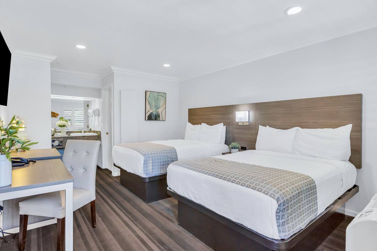 A hotel room with two beds, a desk, and a chair. There is a painting on the wall and a potted plant on the desk.