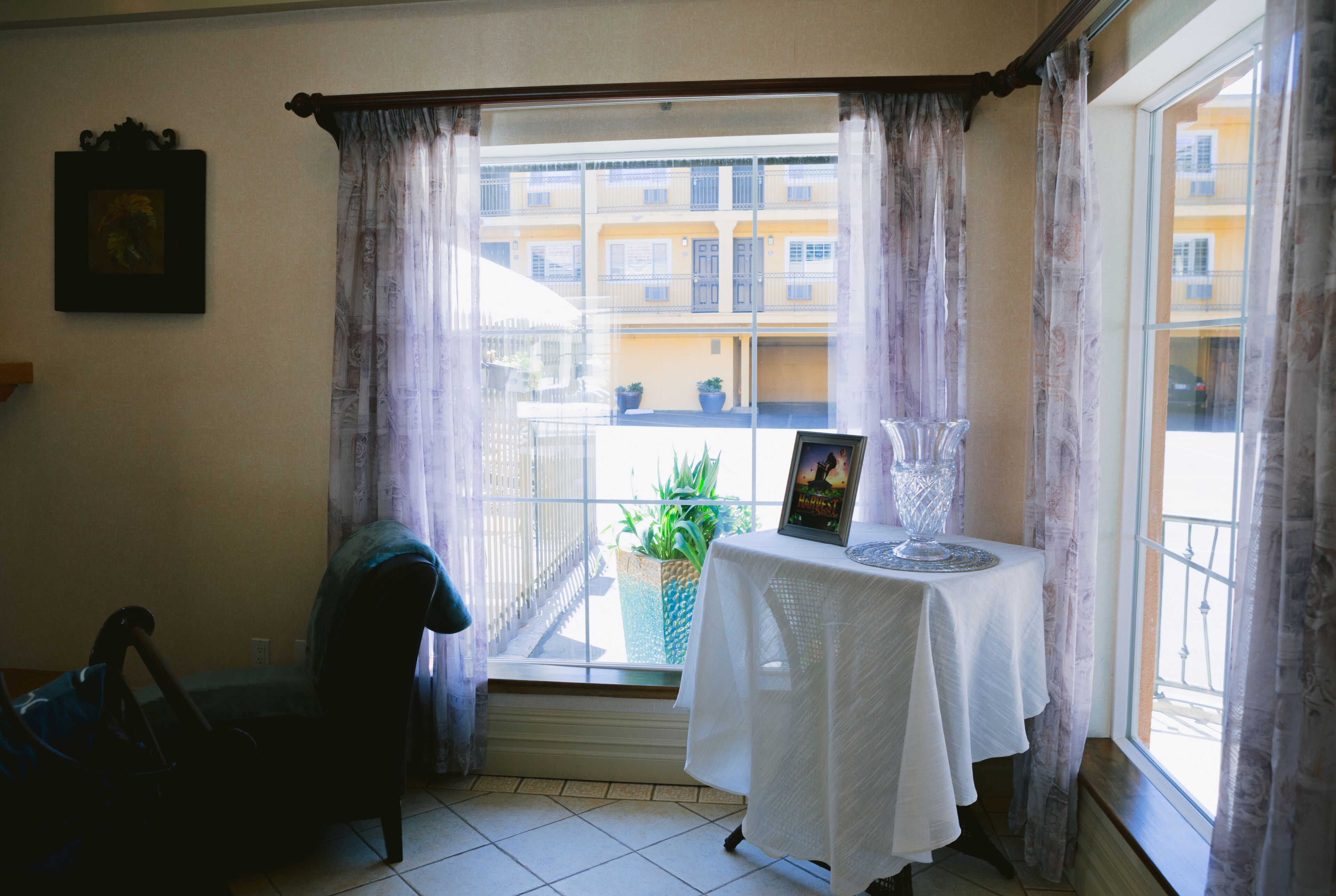 A room with a table set for two, a chair, a window with curtains, and a balcony view.