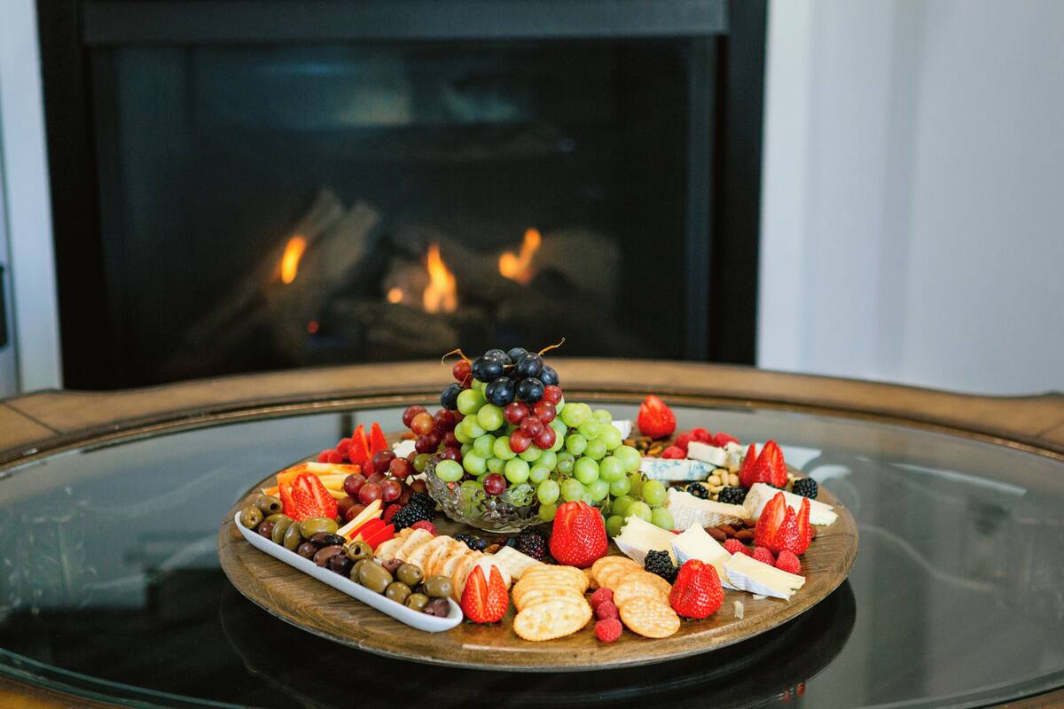 A fruit and cheese platter on a glass table with a fireplace in the background.