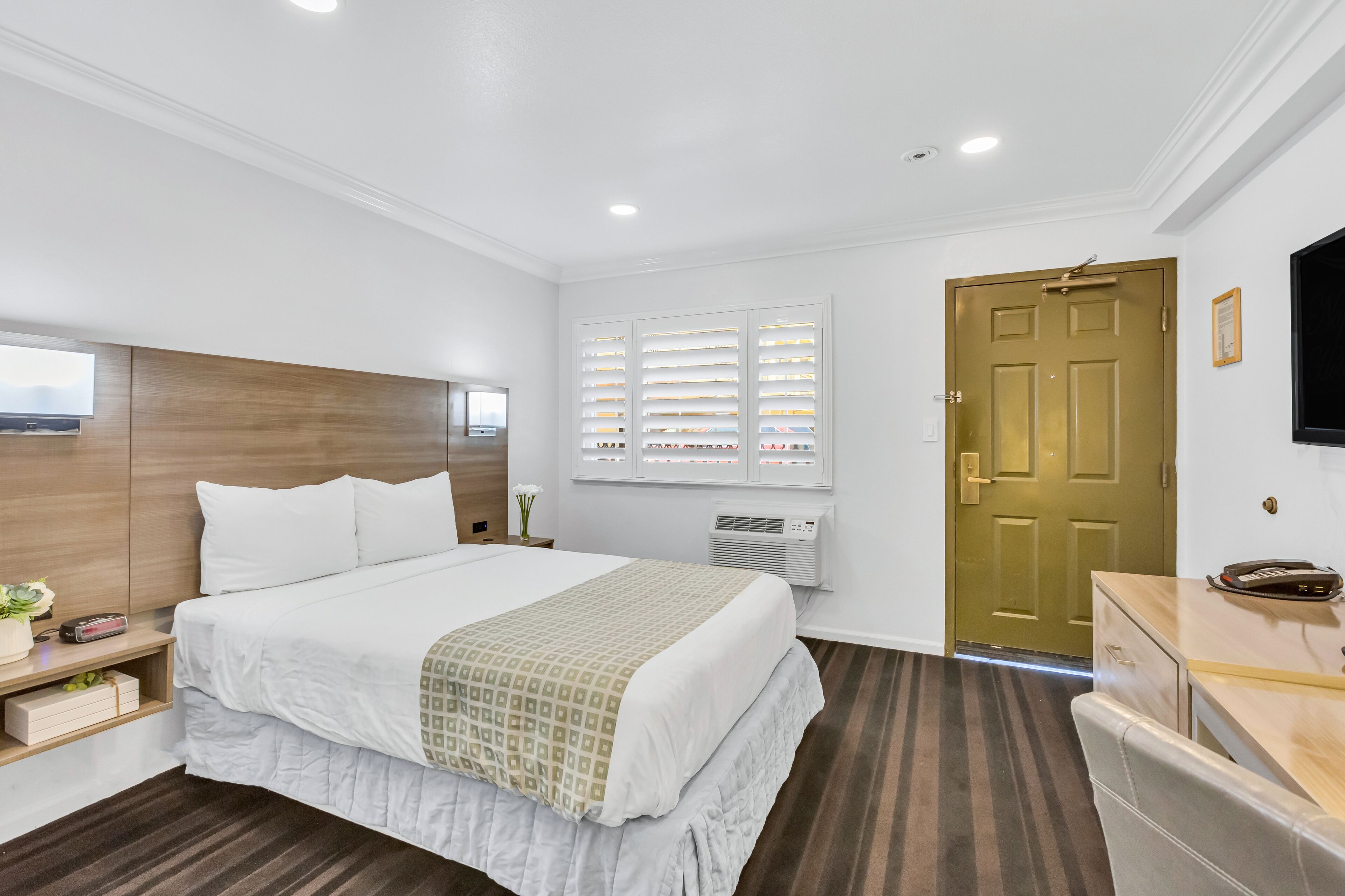 A hotel room with a bed, bedside table, TV, window with blinds, and a door.