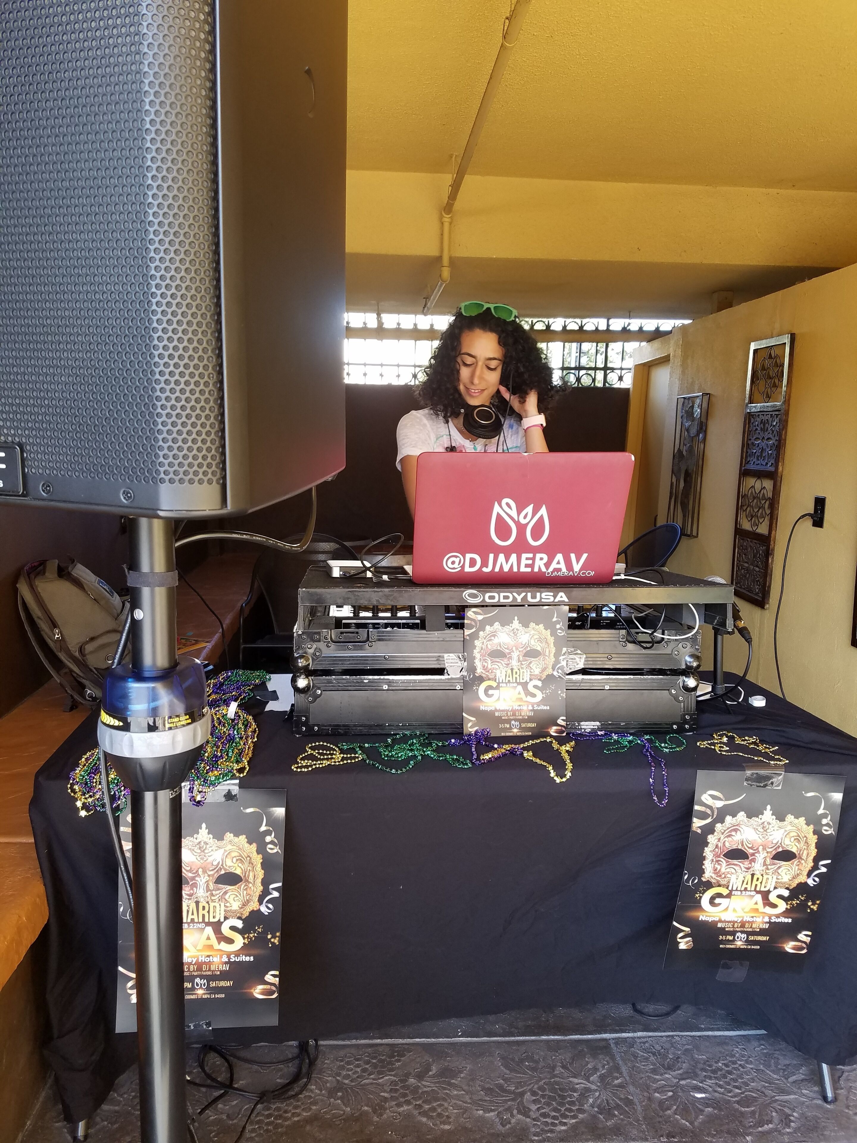 A DJ setup with a laptop, a microphone, and a speaker. There are decorative items on the table, including a Mardi Gras themed poster and colorful beads.
