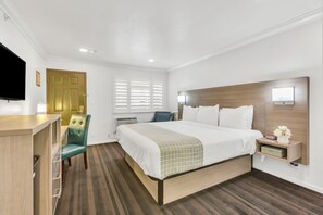 A hotel room with a bed, a desk, a chair, a TV, a window with blinds, and a bedside table with a lamp. - Napa Valley Hotel & Suites (Napa)