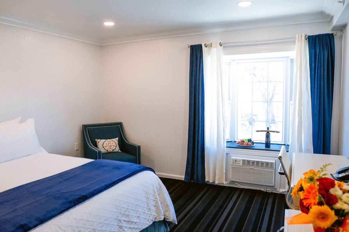 A hotel room with a large bed, a chair, a window with curtains, a desk with a vase of flowers, and an air conditioning unit.