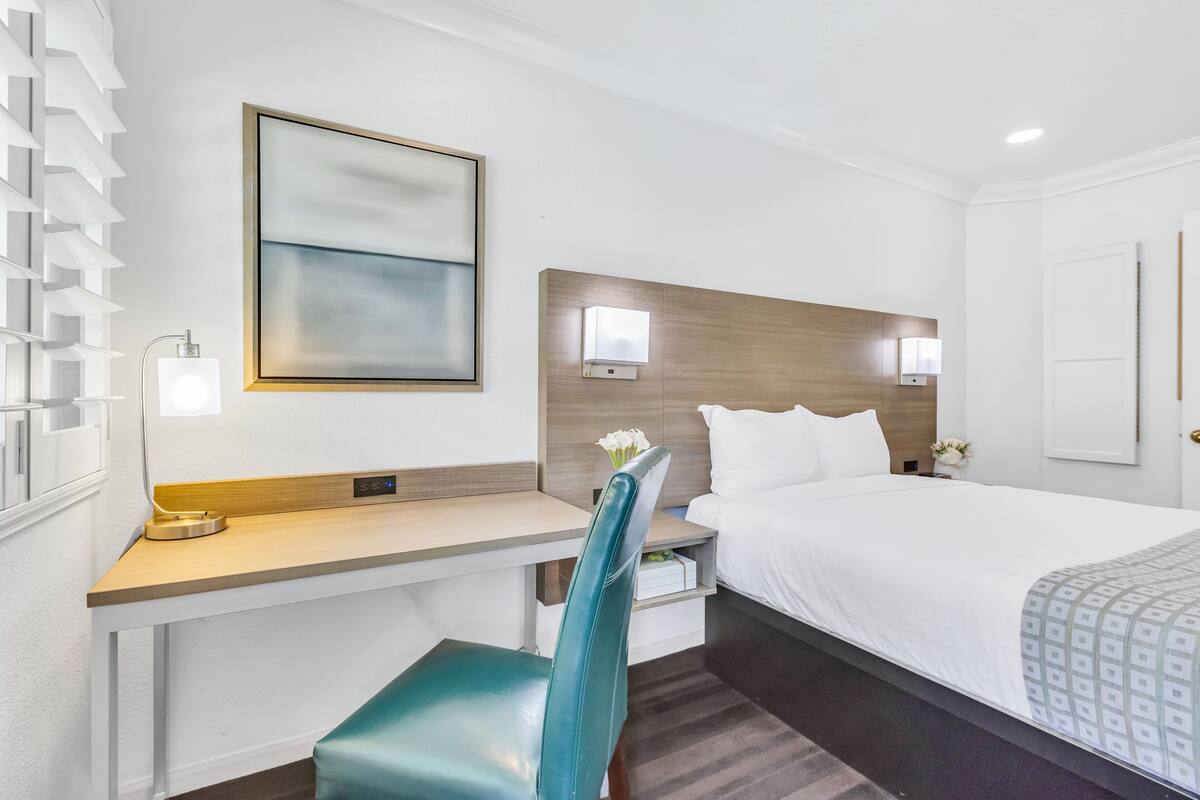 A hotel room with a bed, a desk, a chair, and a bedside table.