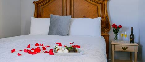 Una cama bien tendida con sábanas blancas, un almohadón gris y pétalos de rosa roja esparcidos sobre la cama. Hay una mesita de noche pequeña con una botella y vasos encima.
