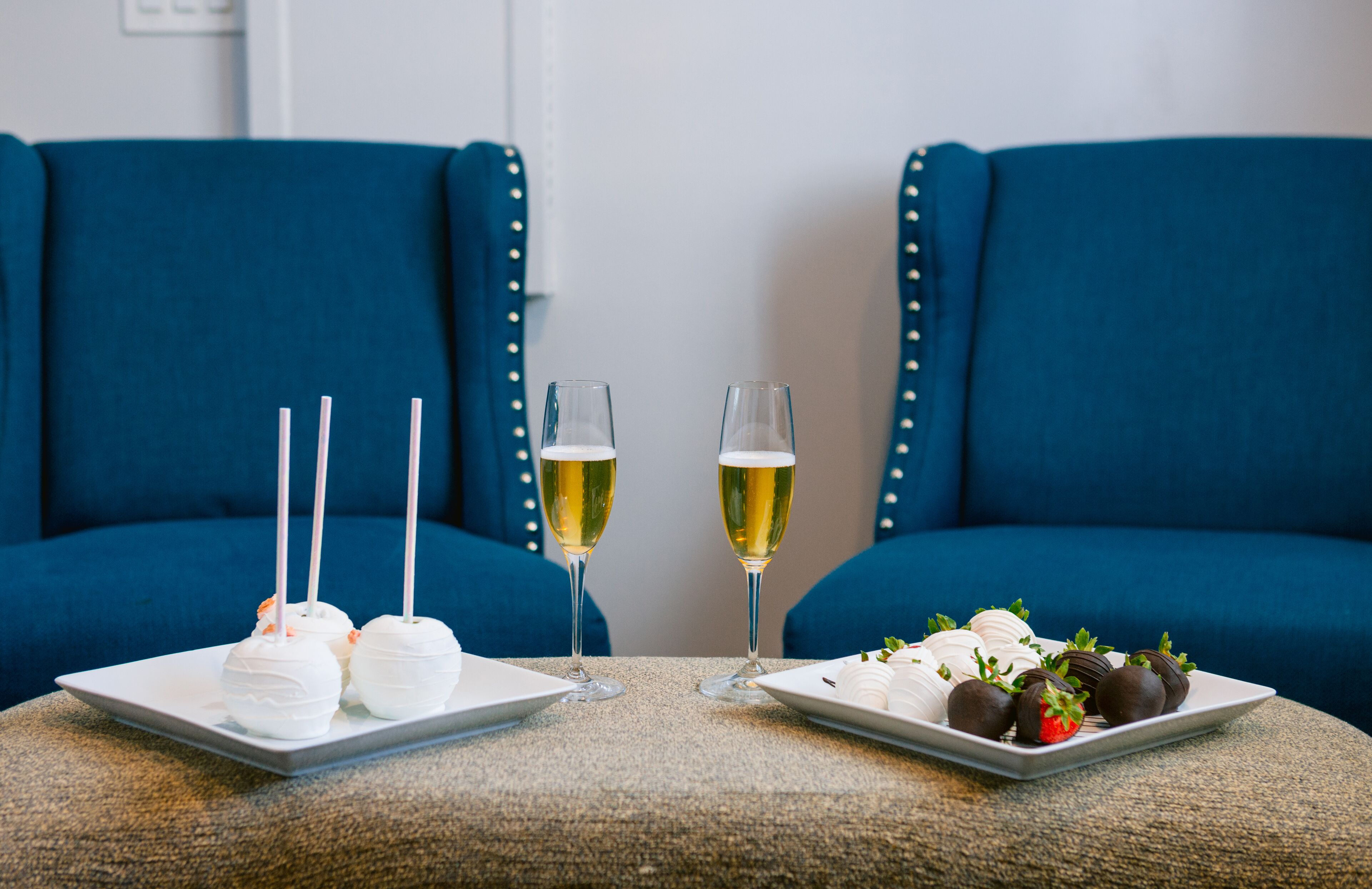 Een tafel met twee vierkante borden, waarvan één witte, bolvormige desserts heeft en de ander aardbeien met chocolade, plus twee glazen witte wijn.