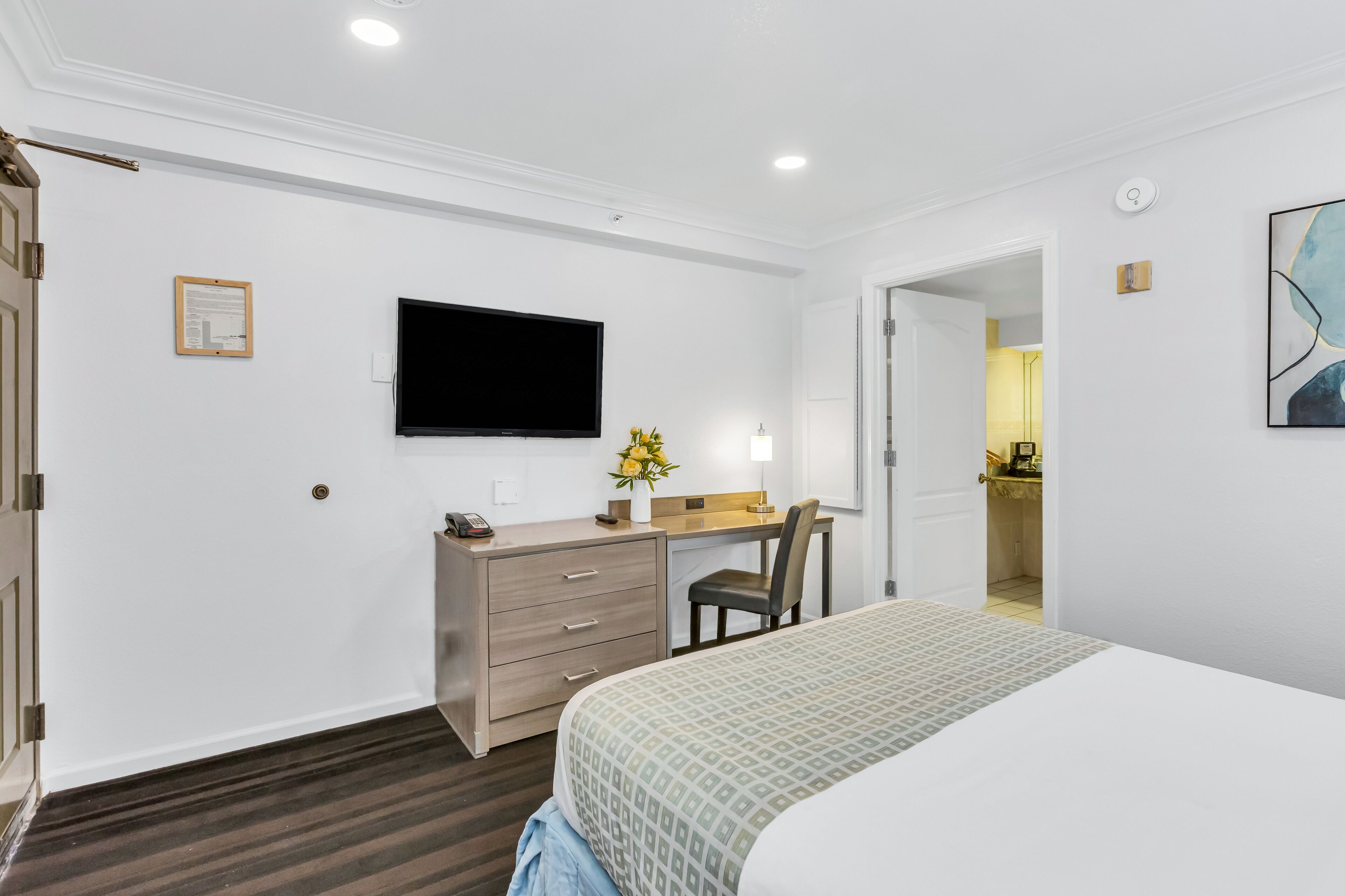 Una habitación de hotel con una cama, un escritorio con una silla, un televisor de pantalla plana en la pared y un pequeño jarrón con flores sobre el escritorio.