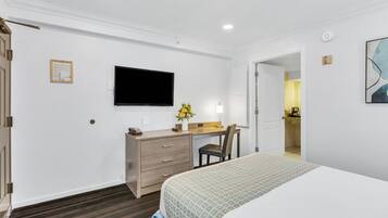 Una habitación de hotel con una cama, un escritorio con una silla, un televisor de pantalla plana en la pared y un pequeño jarrón con flores sobre el escritorio.