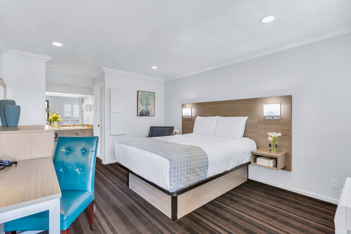 A modern hotel room with a large bed, a desk with a chair, and a small bedside table with a vase of flowers.