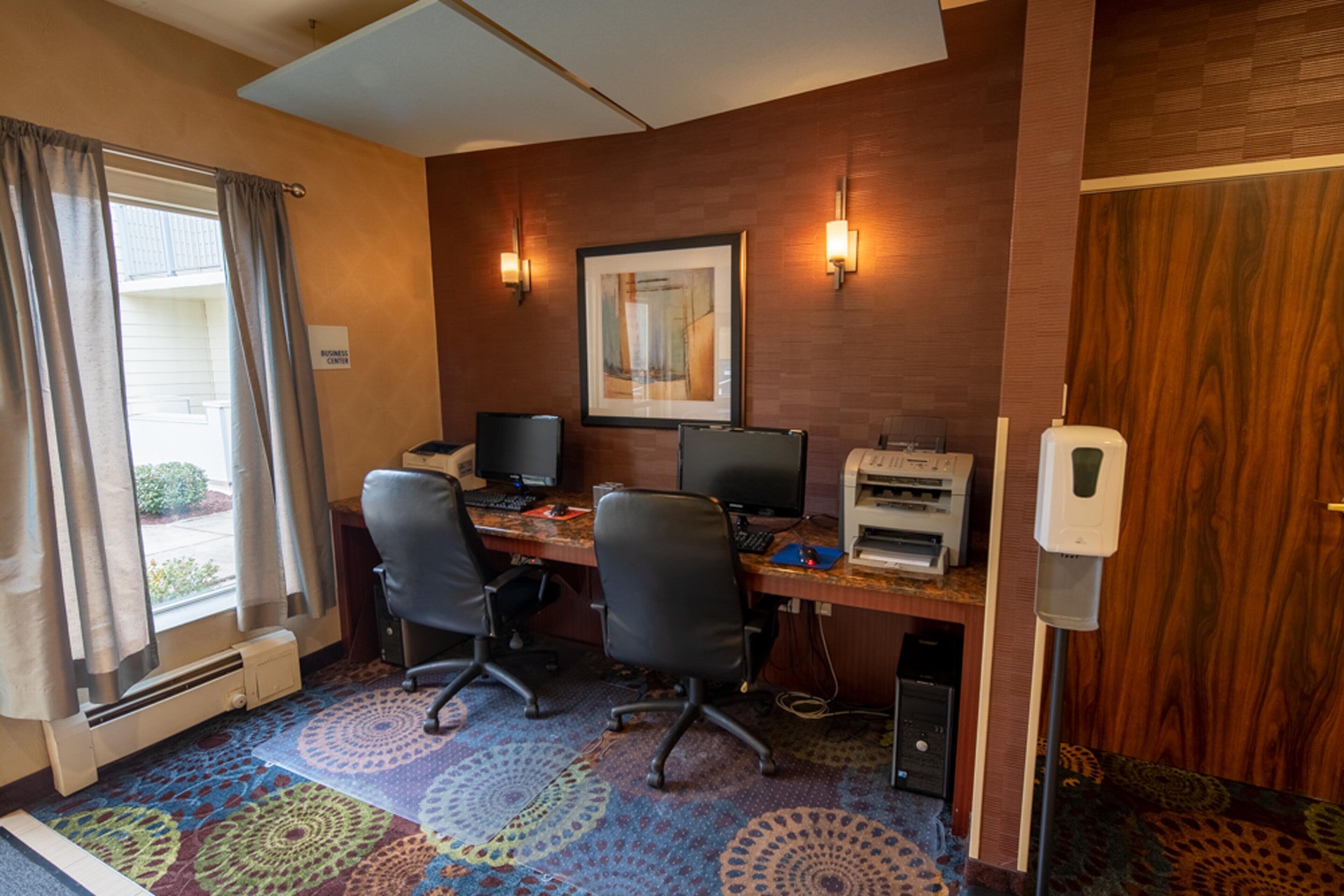 A hotel room with a desk, two chairs, a printer, and a window with curtains.