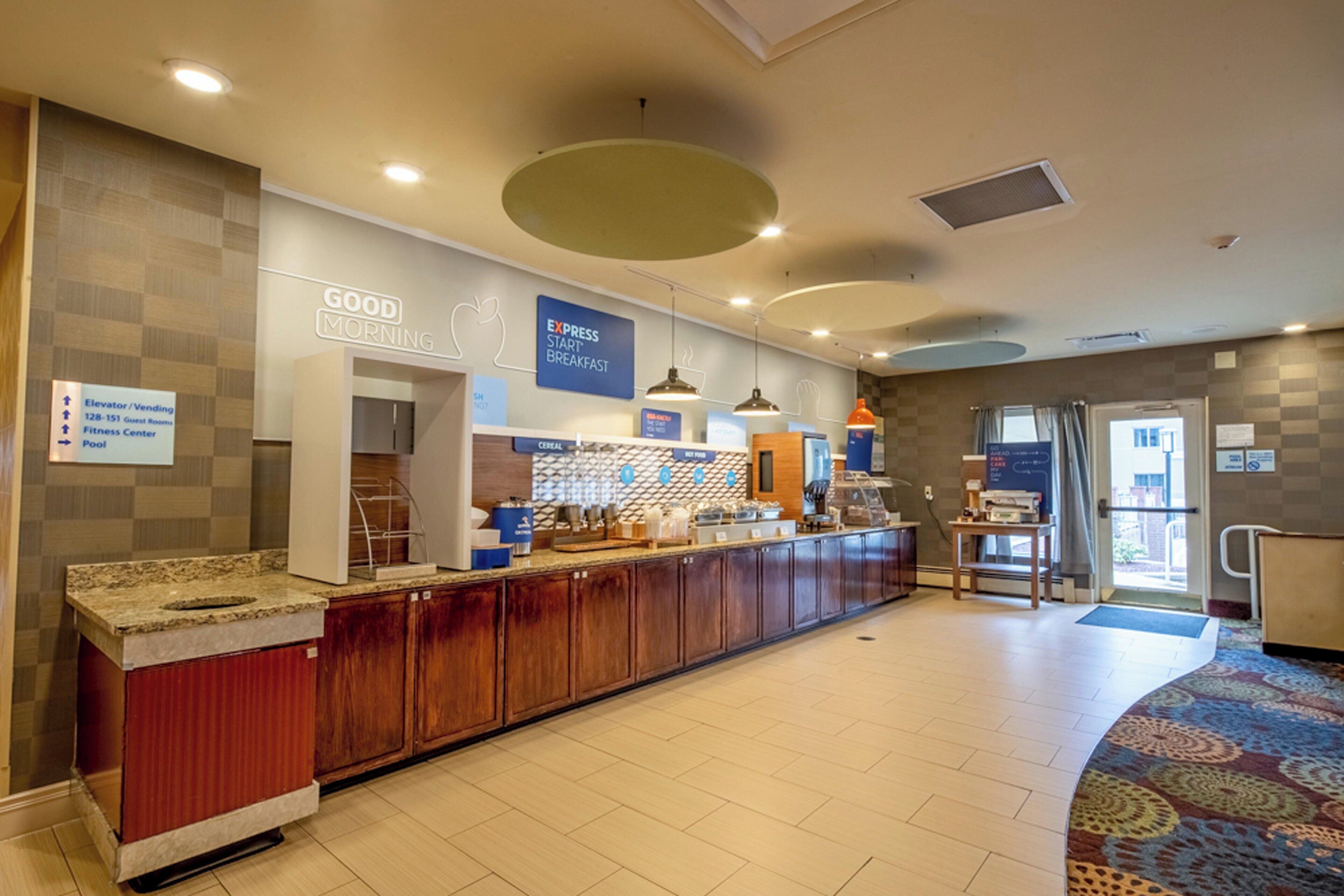 Un vestíbulo de hotel con una mesa de recepción, un letrero que indica desayuno Express Start y señalización que indica la ubicación de servicios como la piscina y el ascensor.