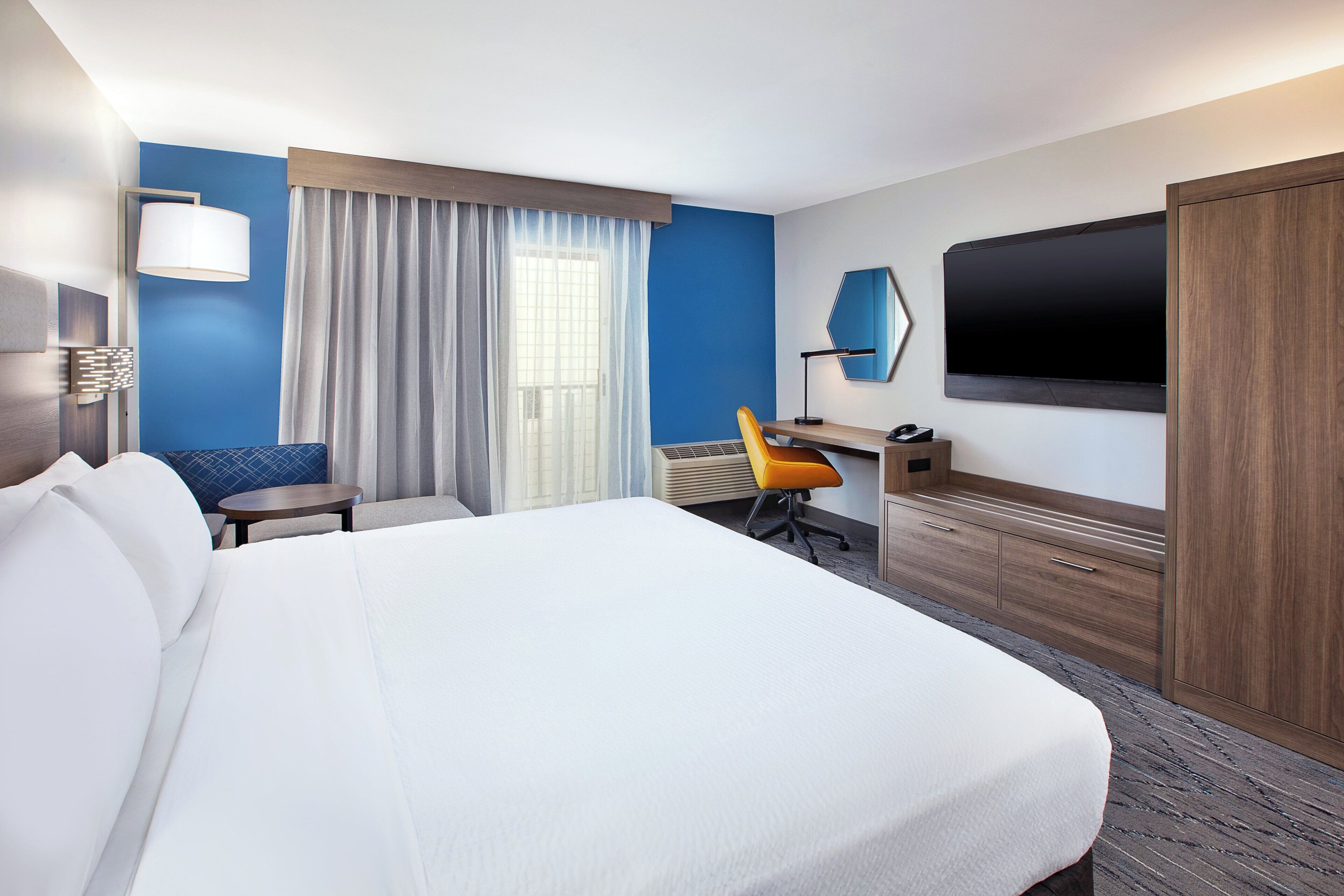 A hotel room with a large bed, a desk with a chair, a flat-screen TV, and a large window with curtains.