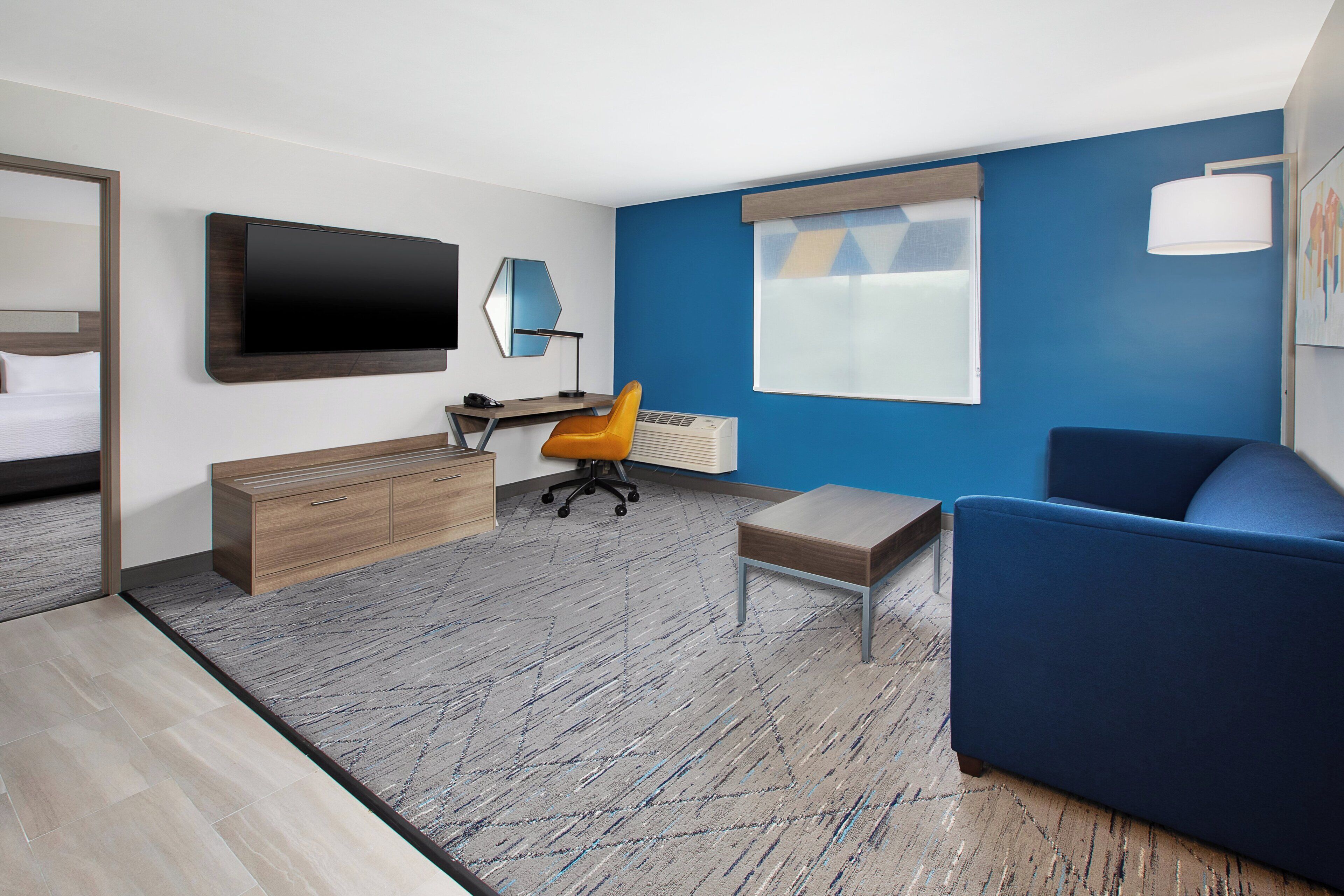 A modern hotel room with a bed, a desk, a blue sofa, and a large TV on the wall.