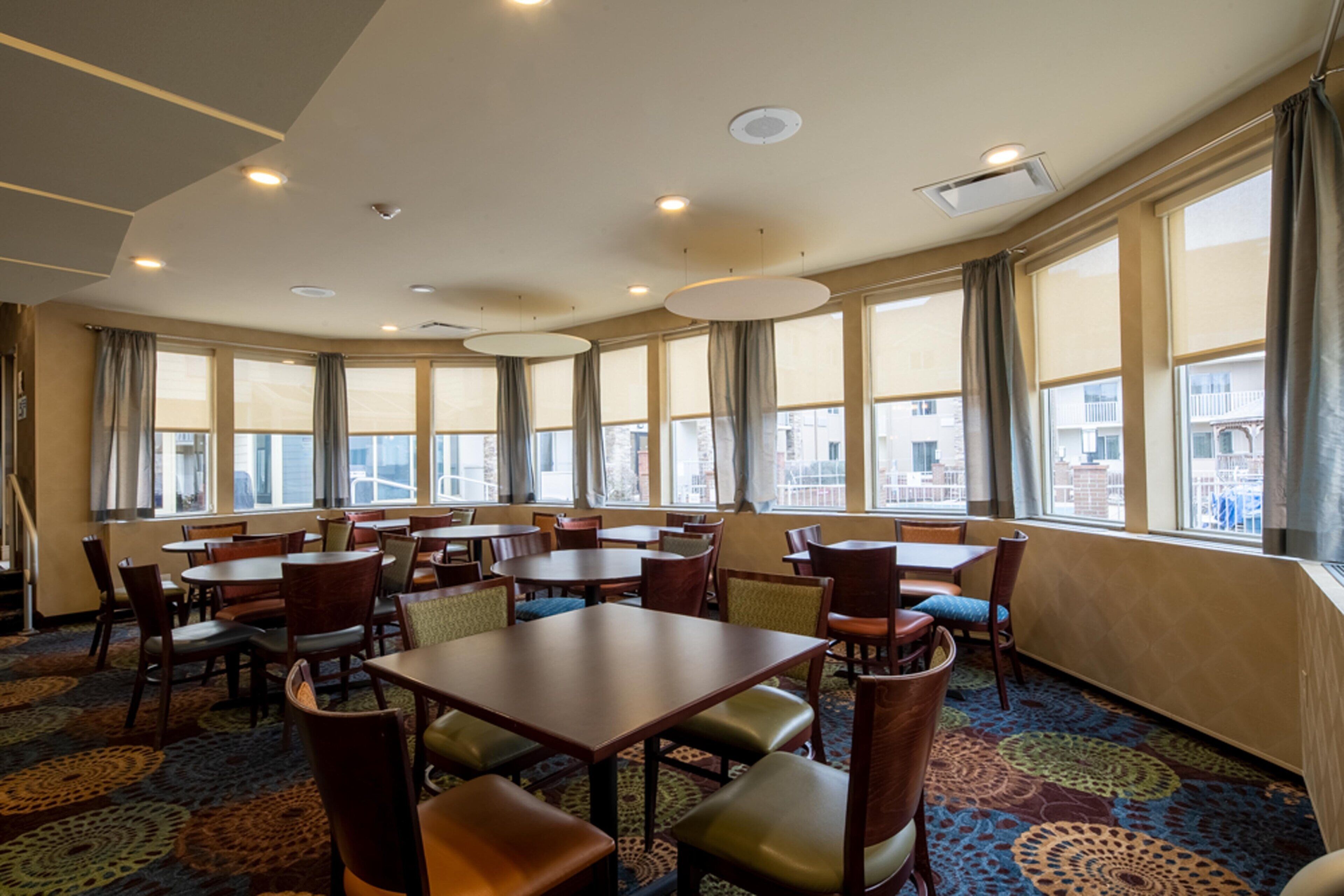 Een restaurantruimte in een hotel met meerdere tafels en stoelen, grote ramen met gordijnen en een kamerbreed tapijt.