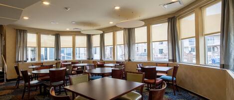 A hotel dining area with multiple tables and chairs, large windows with curtains, and a patterned carpet.