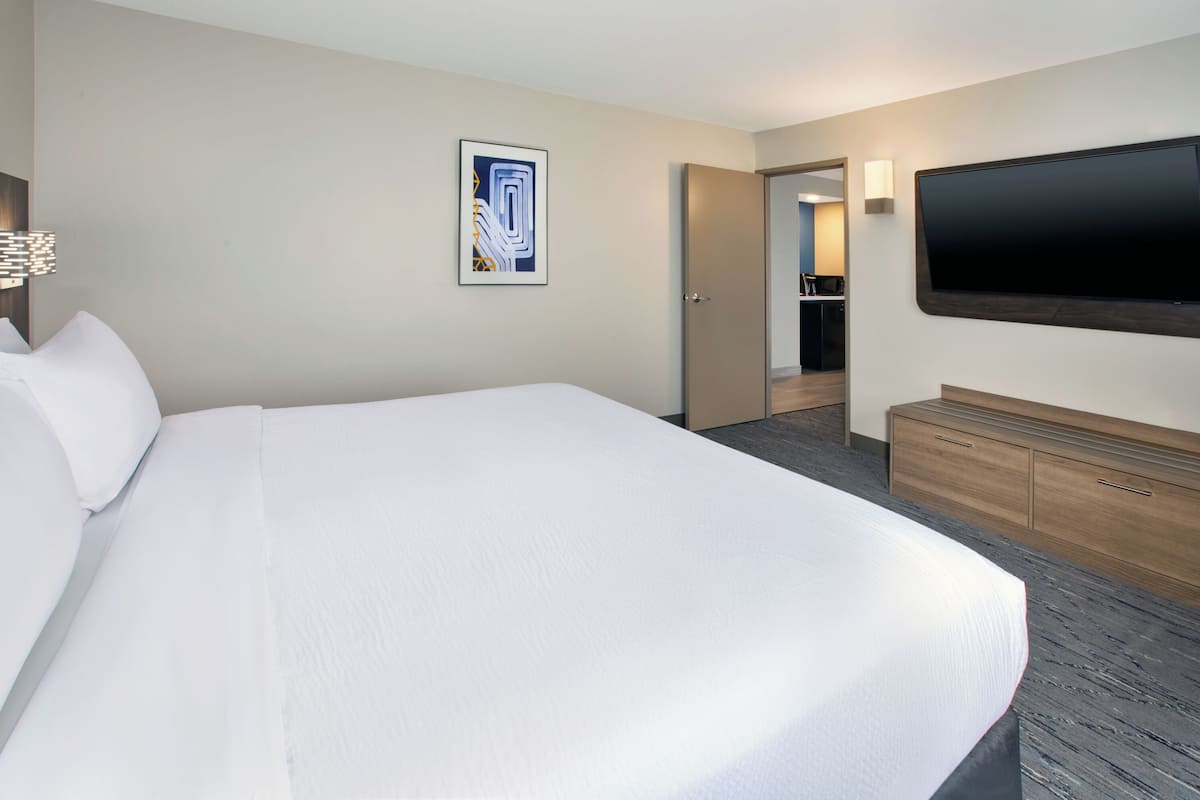 A hotel room with a large bed, a flat-screen TV mounted on the wall, a wooden dresser, and a door leading to a bathroom.