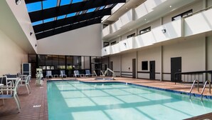 An indoor swimming pool with a clear view of the sky through the ceiling, surrounded by lounge chairs and a brick floor.