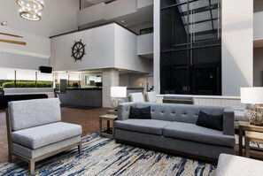 A modern lobby with a grey sofa, armchairs, and a large ship's wheel on the wall. - The Inn on Maritime Bay, an Ascend Collection Hotel (Manitowoc)