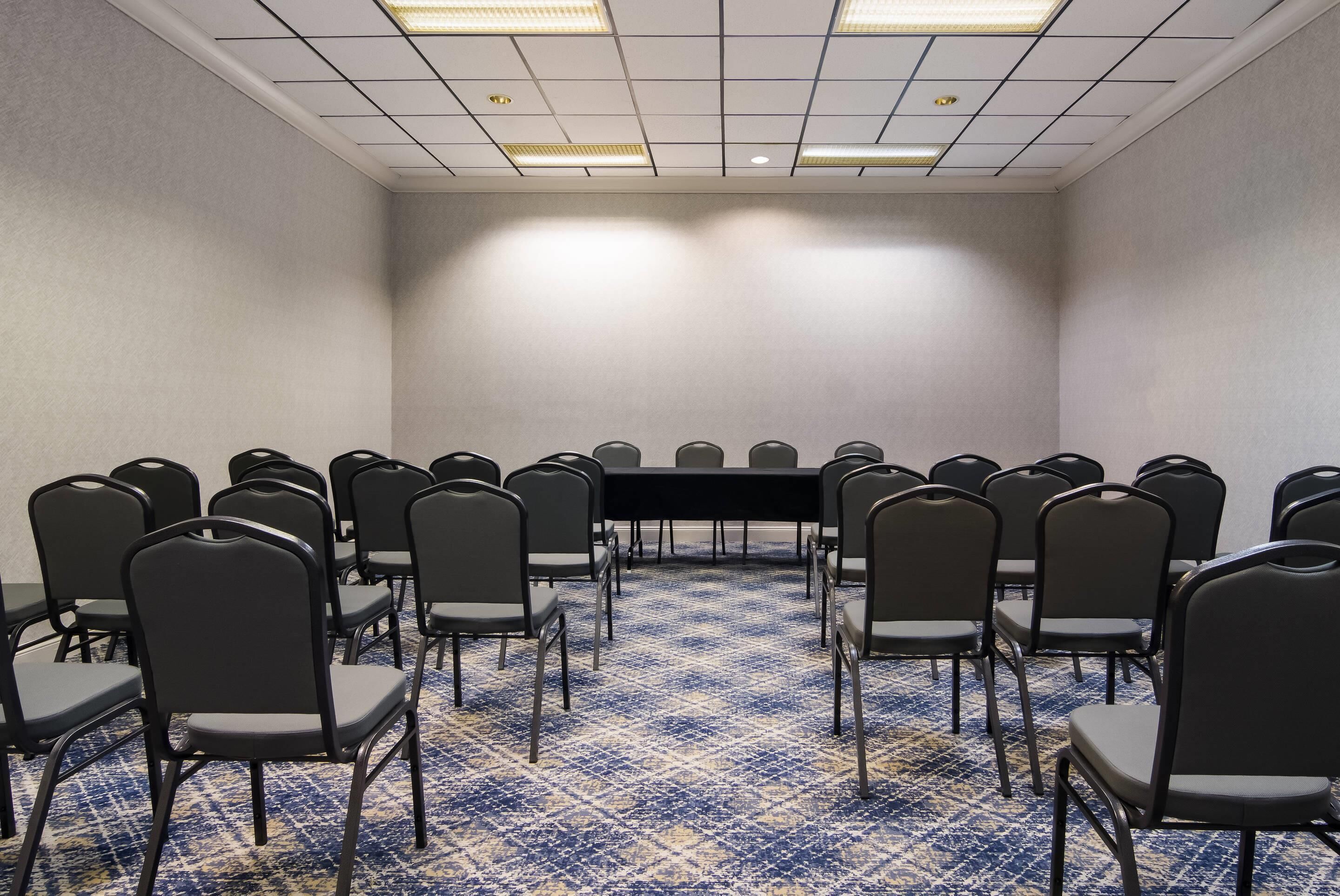 Un salón de conferencias con una larga mesa y filas de sillas, piso alfombrado e iluminación en el techo.