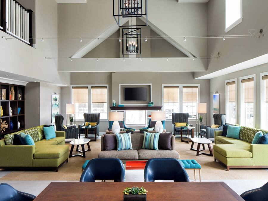 A spacious living room with a high ceiling, a fireplace, and a large flat-screen TV. There are multiple seating areas with sofas and chairs, and a dining table in the center.