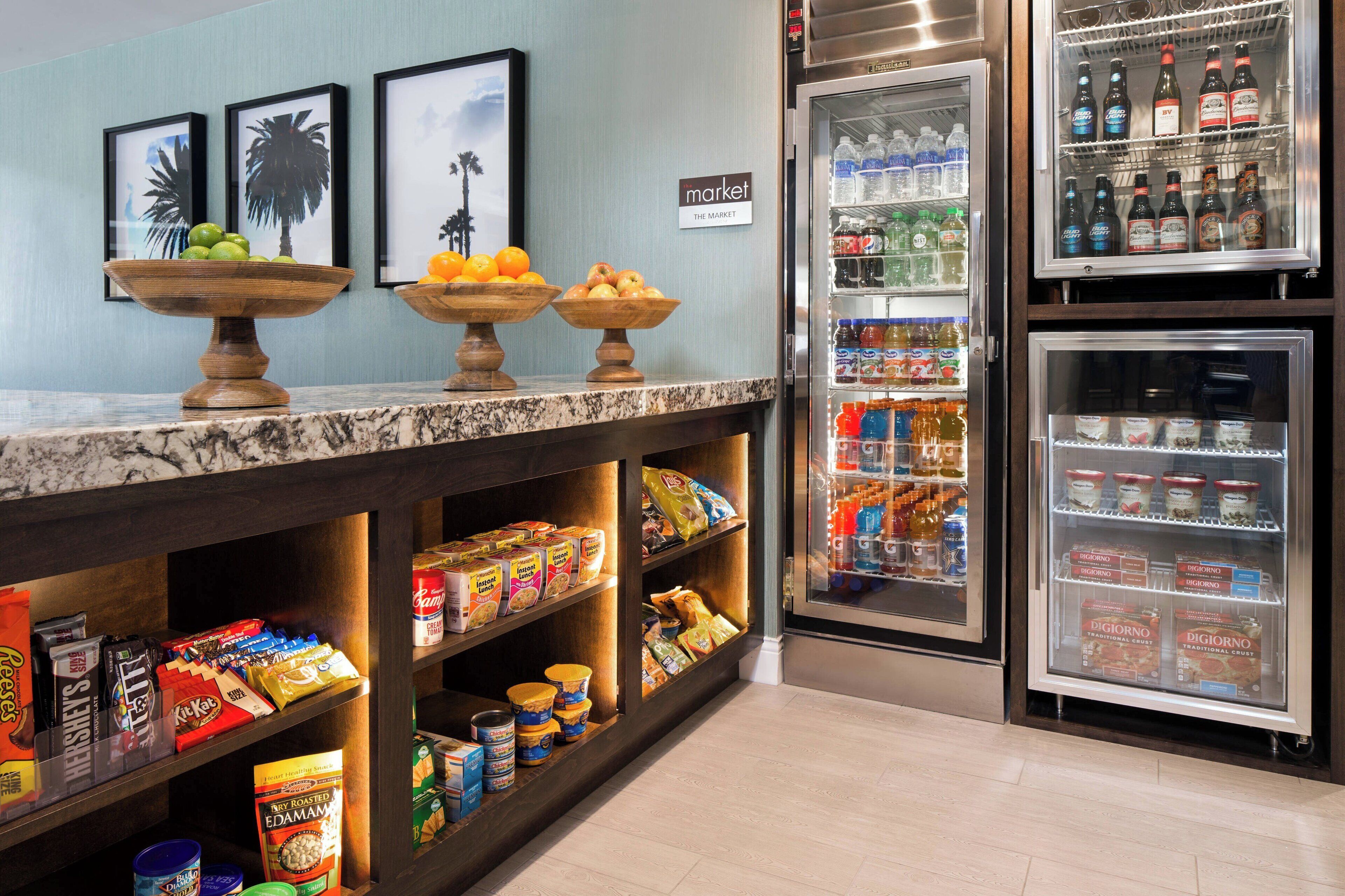 Een hotel receptie met een marmeren balie, twee houten schalen vol fruit, een koelkast gevuld met drankjes, en planken met snacks en toevoegingen.