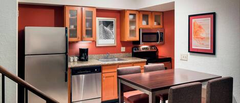 A compact kitchen with a refrigerator, microwave, and dishwasher. There is a dining table with chairs and a framed picture on the wall.