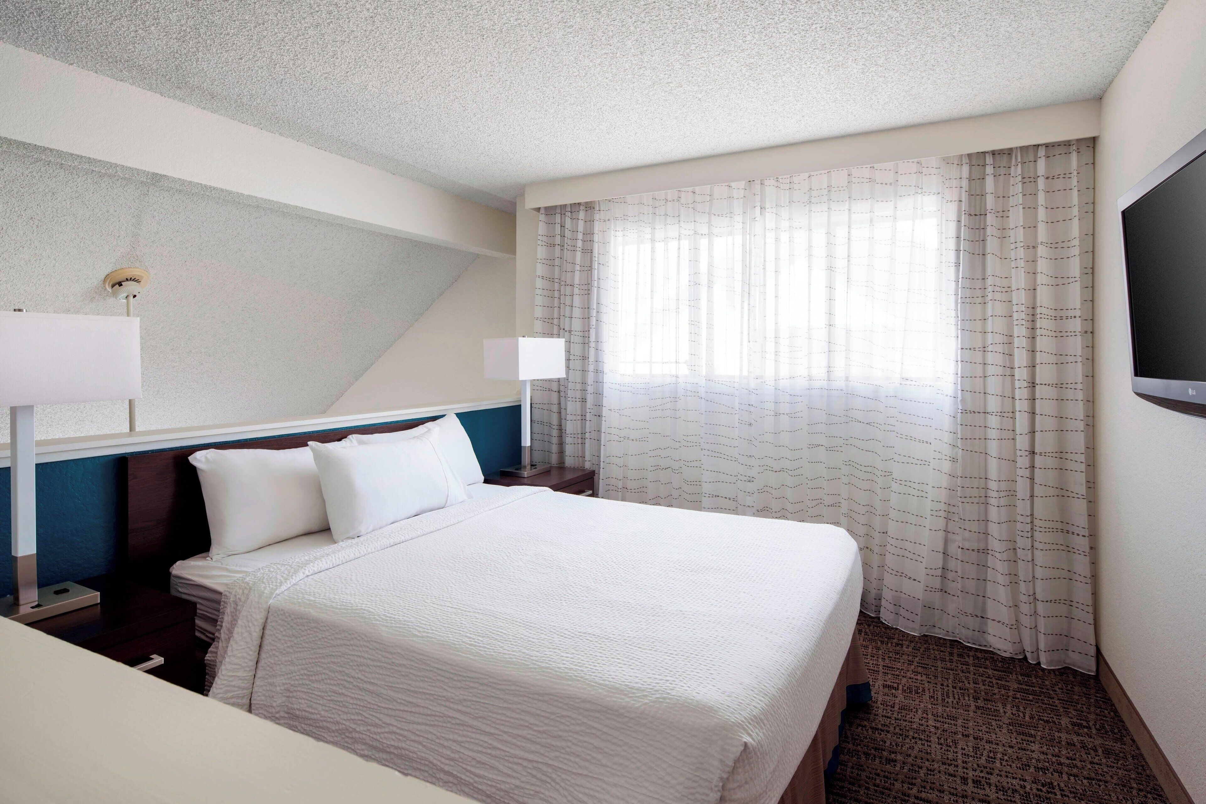 A hotel room with a large bed, two bedside tables, a flat-screen TV, and a window with sheer curtains.