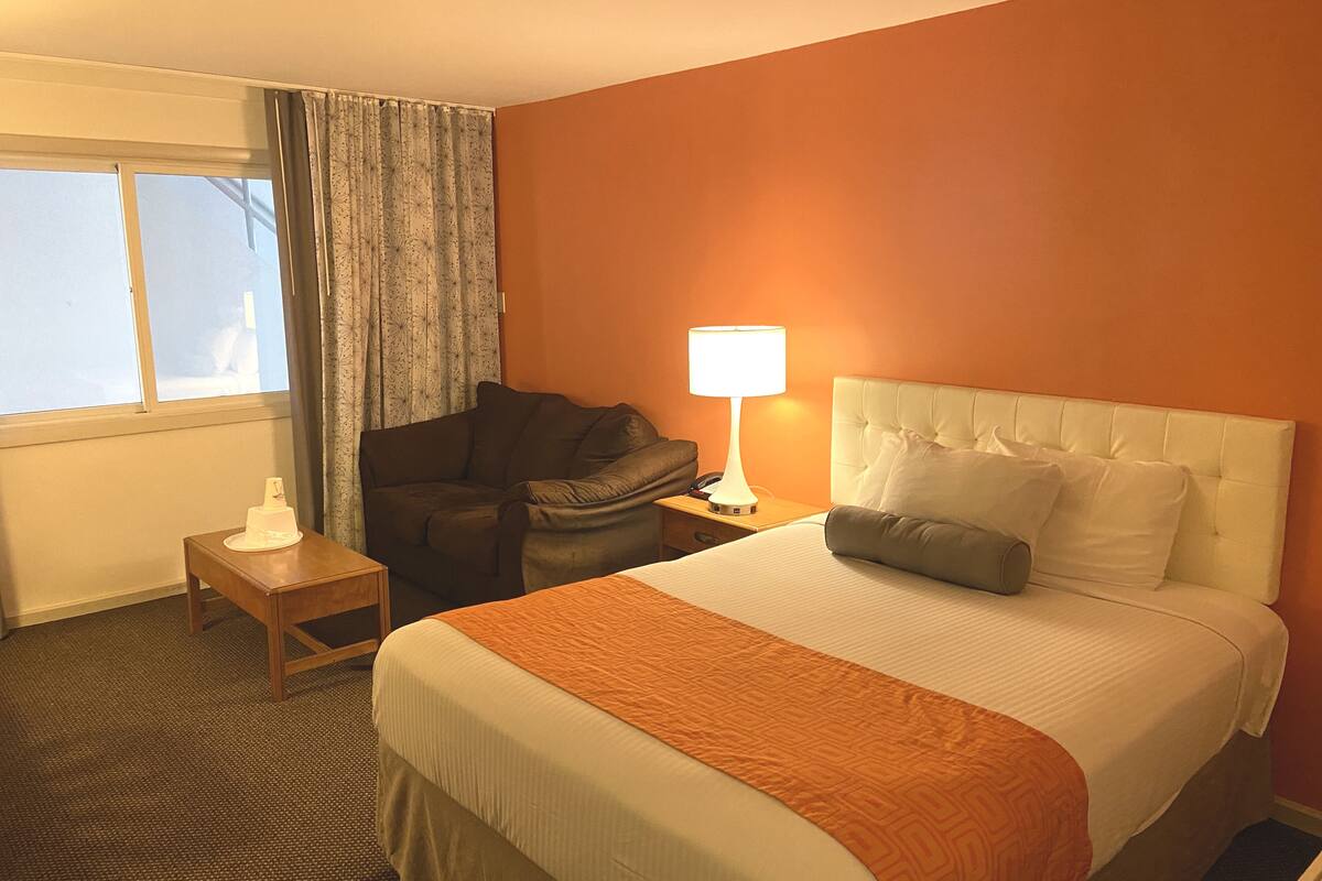 A hotel room with a bed, a nightstand, a lamp, a chair, and a window with curtains.