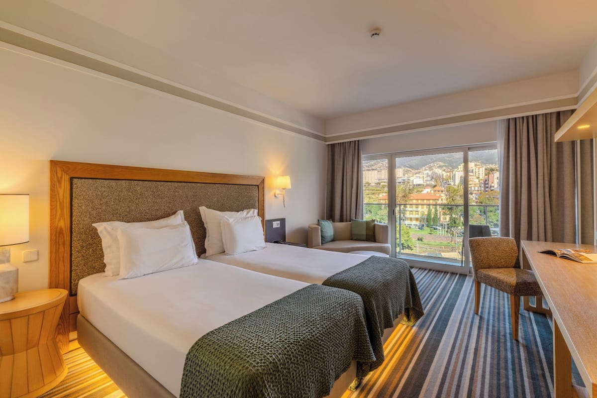 Habitación de hotel con dos camas, un escritorio, una silla, un sofá y un amplio ventanal con vista a la ciudad.