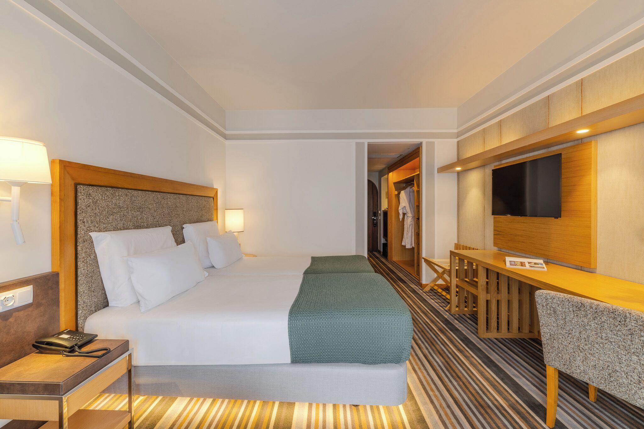 A hotel room with a bed, bedside table, lamp, TV, and a small desk area.