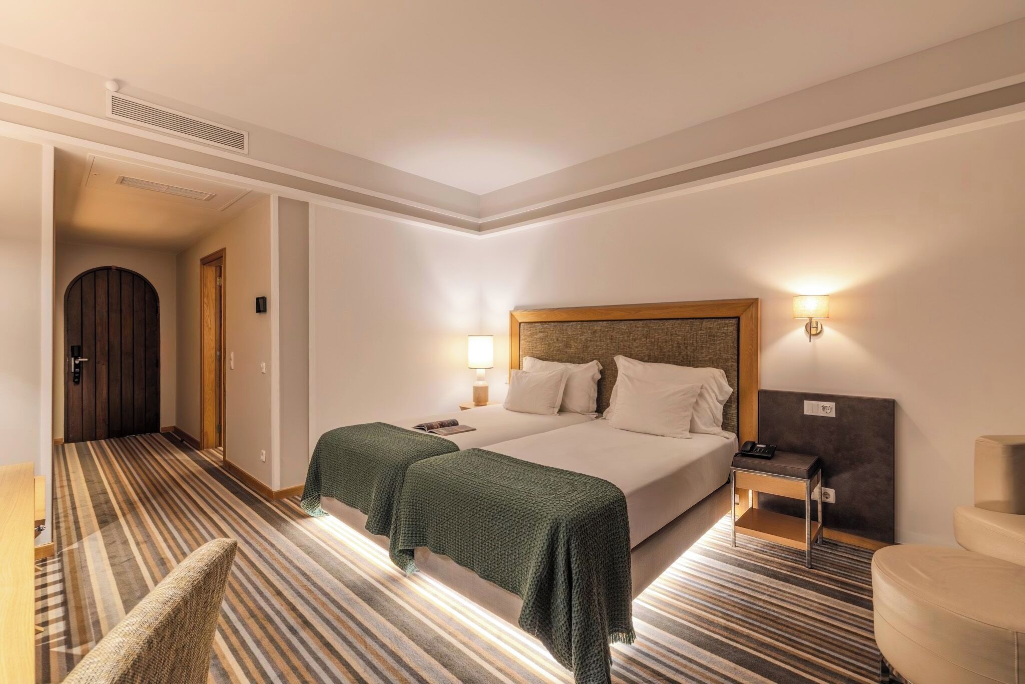 Una habitación de hotel con una cama, dos mesitas de noche, una silla, una lámpara y una puerta que da a un pasillo.