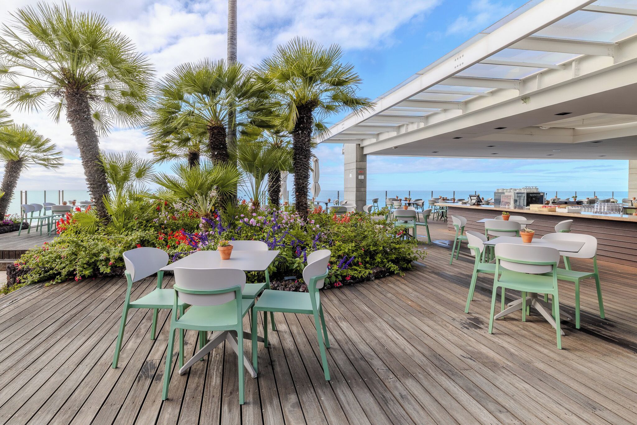An outdoor dining area with wooden flooring, green chairs, and tables, surrounded by lush greenery and palm trees, overlooking a body of water.