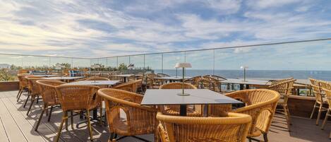 A rooftop terrace with wicker chairs and tables, overlooking a coastal view.