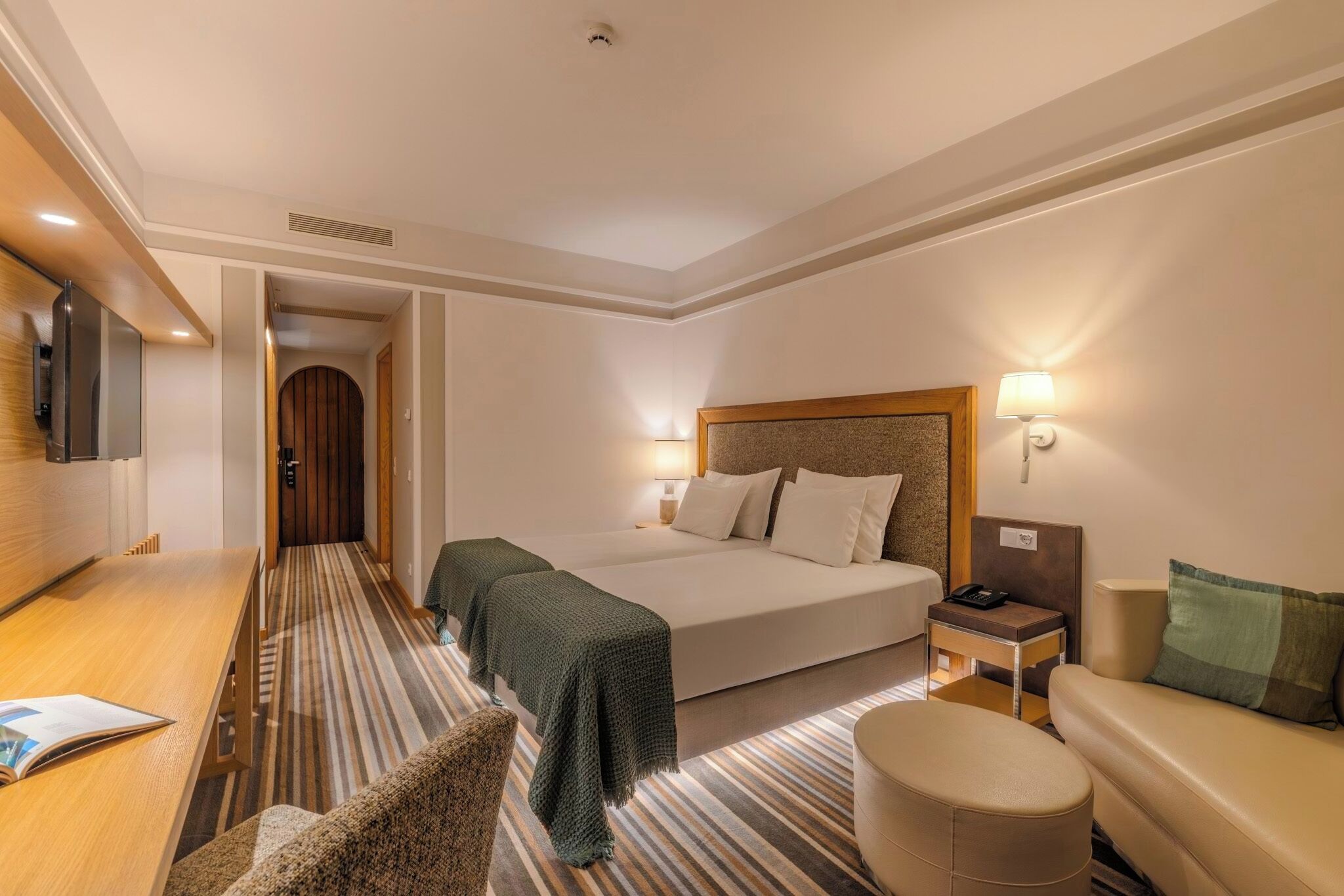 A hotel room with a bed, bedside table, lamp, and a view of a hallway.