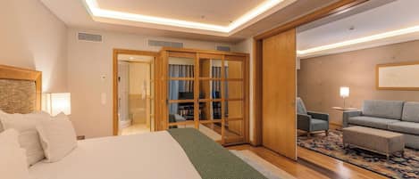 Habitación de hotel con cama, zona de estar con sofá y sillones, armario de madera y puerta corredera que da a un baño.