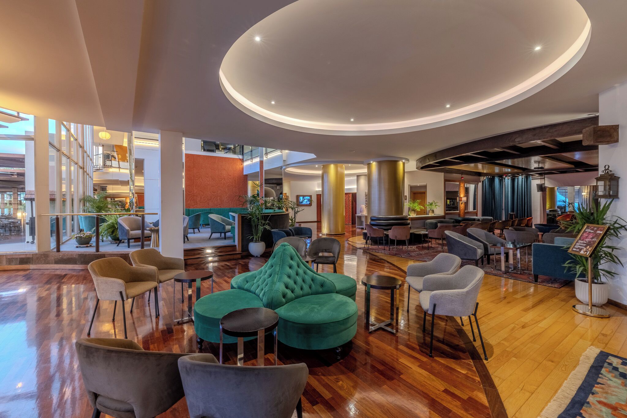 A spacious lobby with a central seating area featuring a green sofa and multiple chairs, a bar area with stools, and a large open space with a wooden floor.