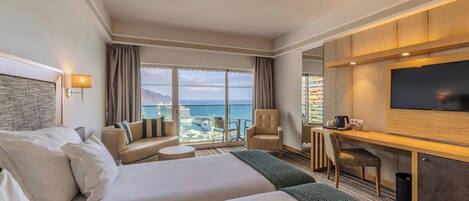 A hotel room with two beds, a desk, a TV, and a view of the ocean.